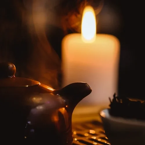 Warm evening tea arrangement featuring a ceramic kyusu teapot, handblown glass cup, beeswax candles, and soft blanket creating a cosy quiet night atmosphere